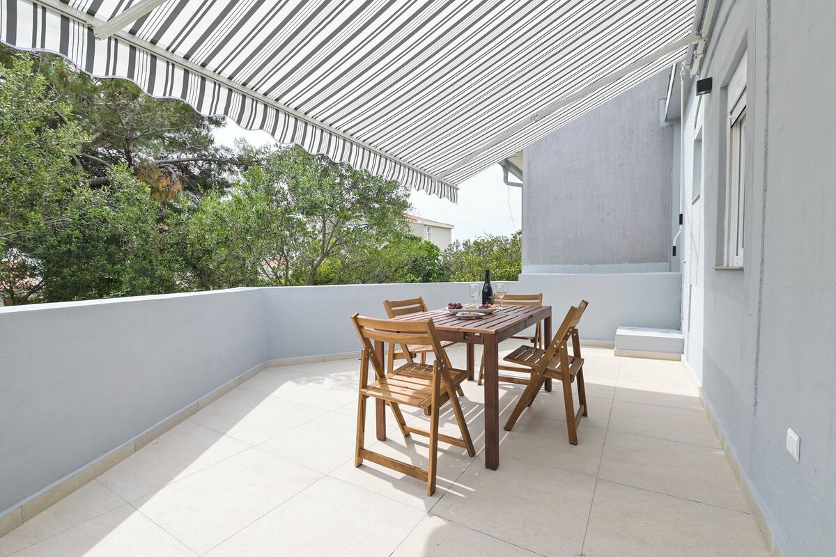 A covered outdoor patio with a wooden table and chairs, surrounded by a low white wall and greenery.