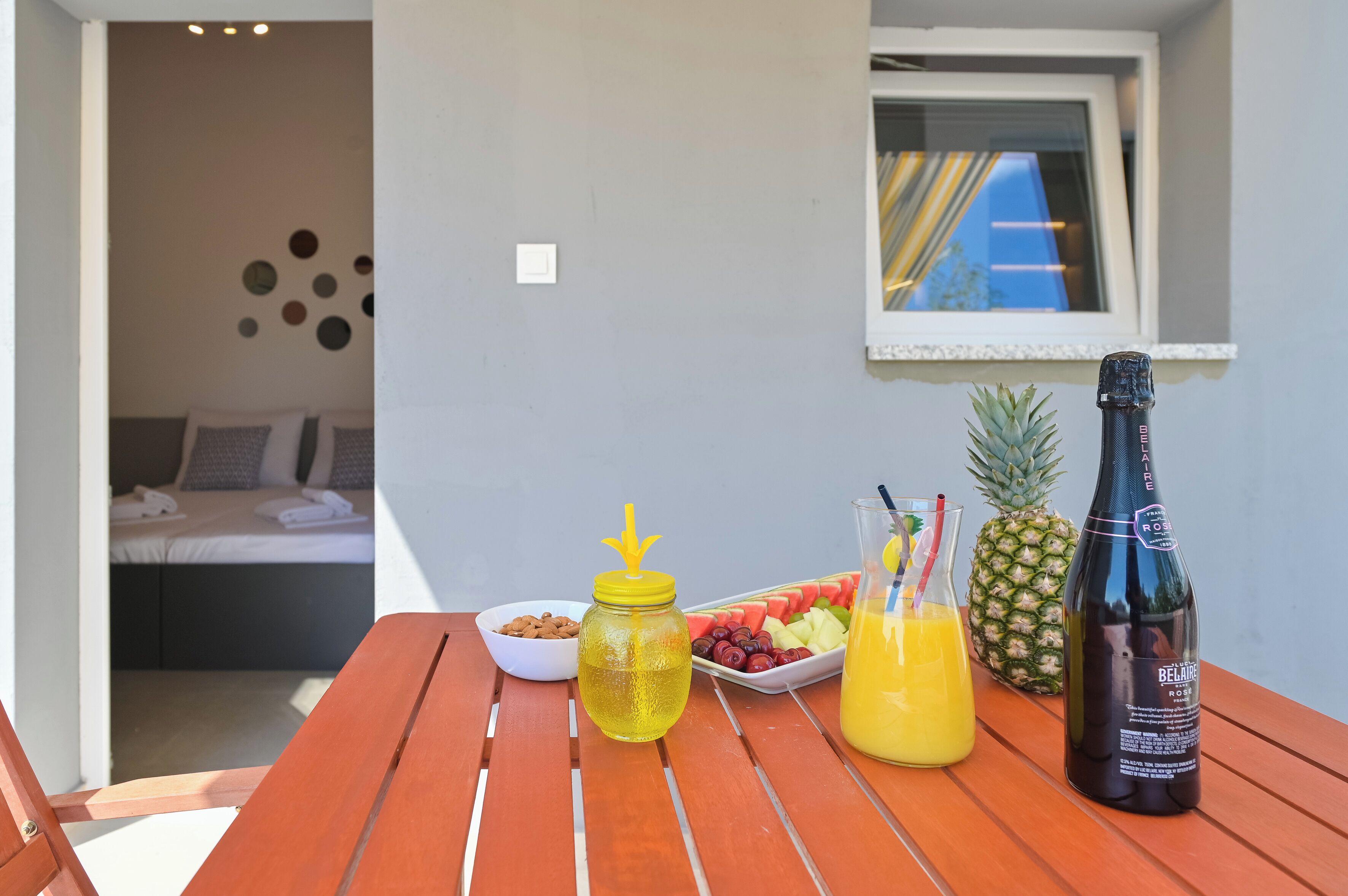 A wooden table with a bottle of rosé, a glass of orange juice, a bowl of nuts, a fruit platter, and a pineapple.