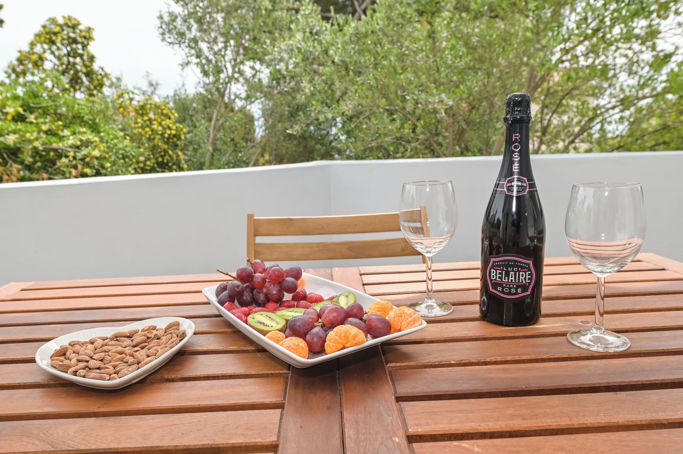 A table with a bottle of Belair Rose wine, two glasses, a plate of assorted fruits, and a bowl of almonds.