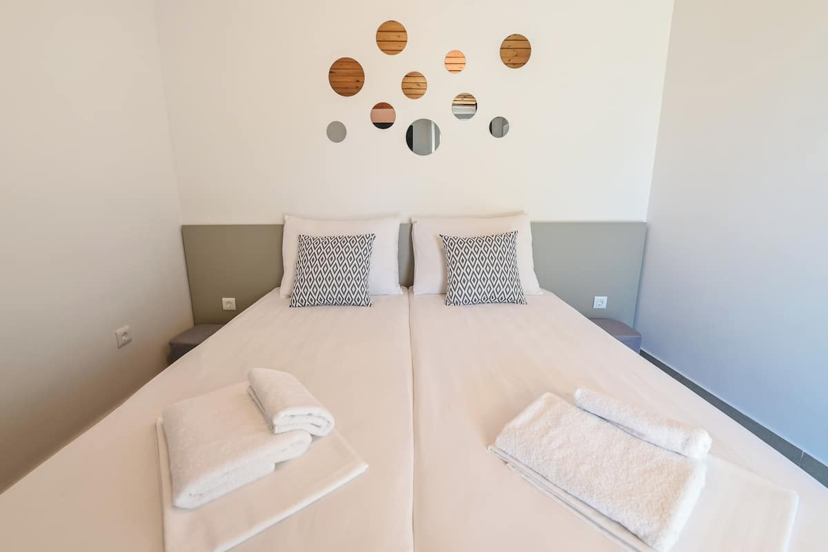 A double bed with white linens and patterned pillows, a wall-mounted mirror, and a towel on the bed.