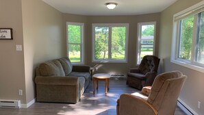 Ein Wohnzimmer mit Ecksofa, einem Sessel und einem kleinen Tisch. Drei große Fenster bieten Blick auf eine grüne Landschaft.