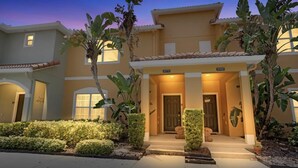 Una casa a due piani con un ingresso ben illuminato, un piccolo portico e un giardino curato con diverse piante.