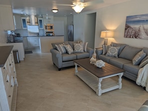 Una sala de estar moderna con un sofá gris, una mesa de centro de madera y una cocina al fondo.