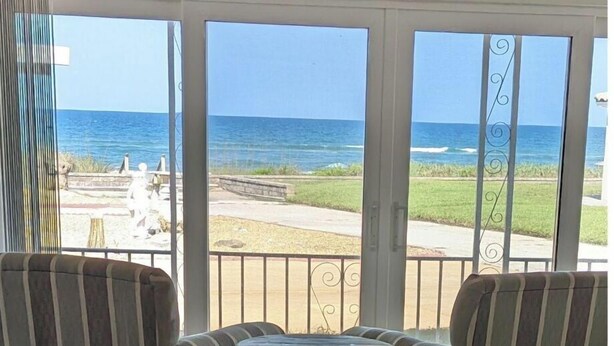 Una stanza con due poltrone a righe e un tavolino rotondo, con vista su una spiaggia, l'oceano e una zona verde.