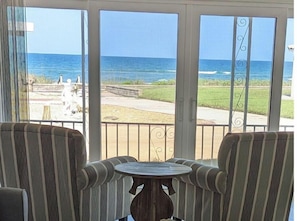 Una habitación con dos sillones a rayas y una pequeña mesa redonda, con vista a una playa que incluye el océano y una zona cubierta de césped.
