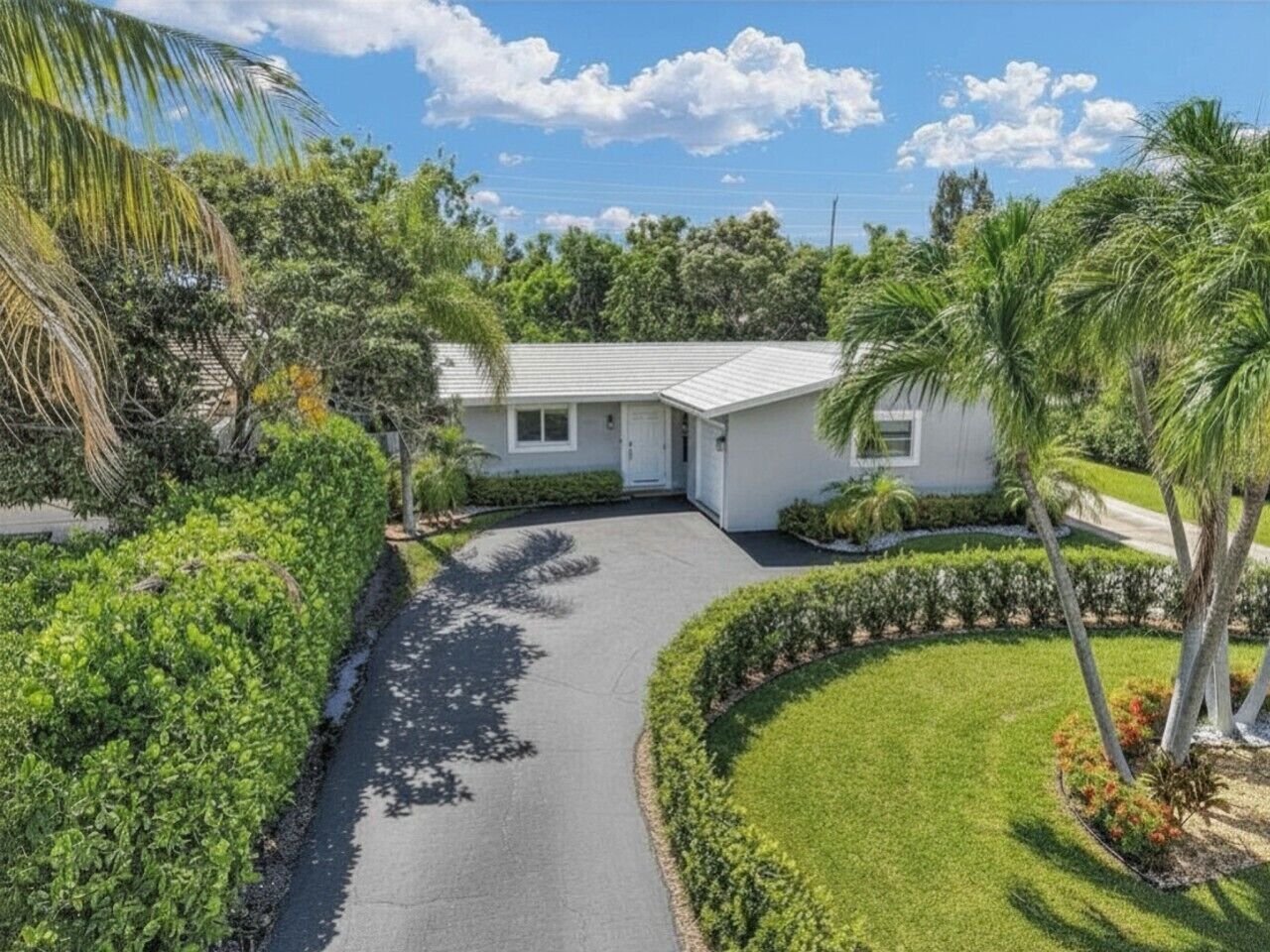 A residential property with a paved driveway, a white house, and a well-maintained lawn with palm trees.