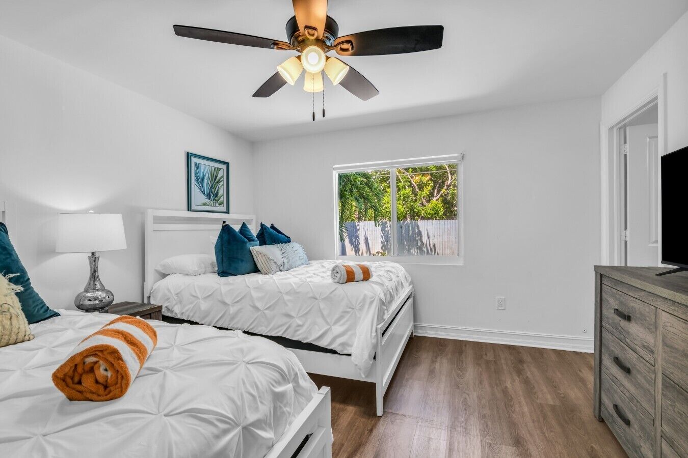 A bedroom with two beds, a ceiling fan, a window with a view of greenery, and a wooden dresser.