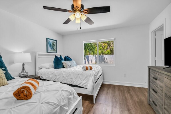 A bedroom with two beds, a ceiling fan, a window with a view of greenery, and a wooden dresser.
