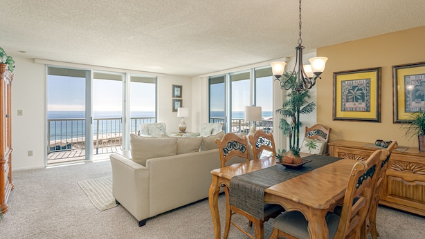 A spacious living room with a balcony overlooking the ocean, a dining area with wooden furniture, and a view of the beach.