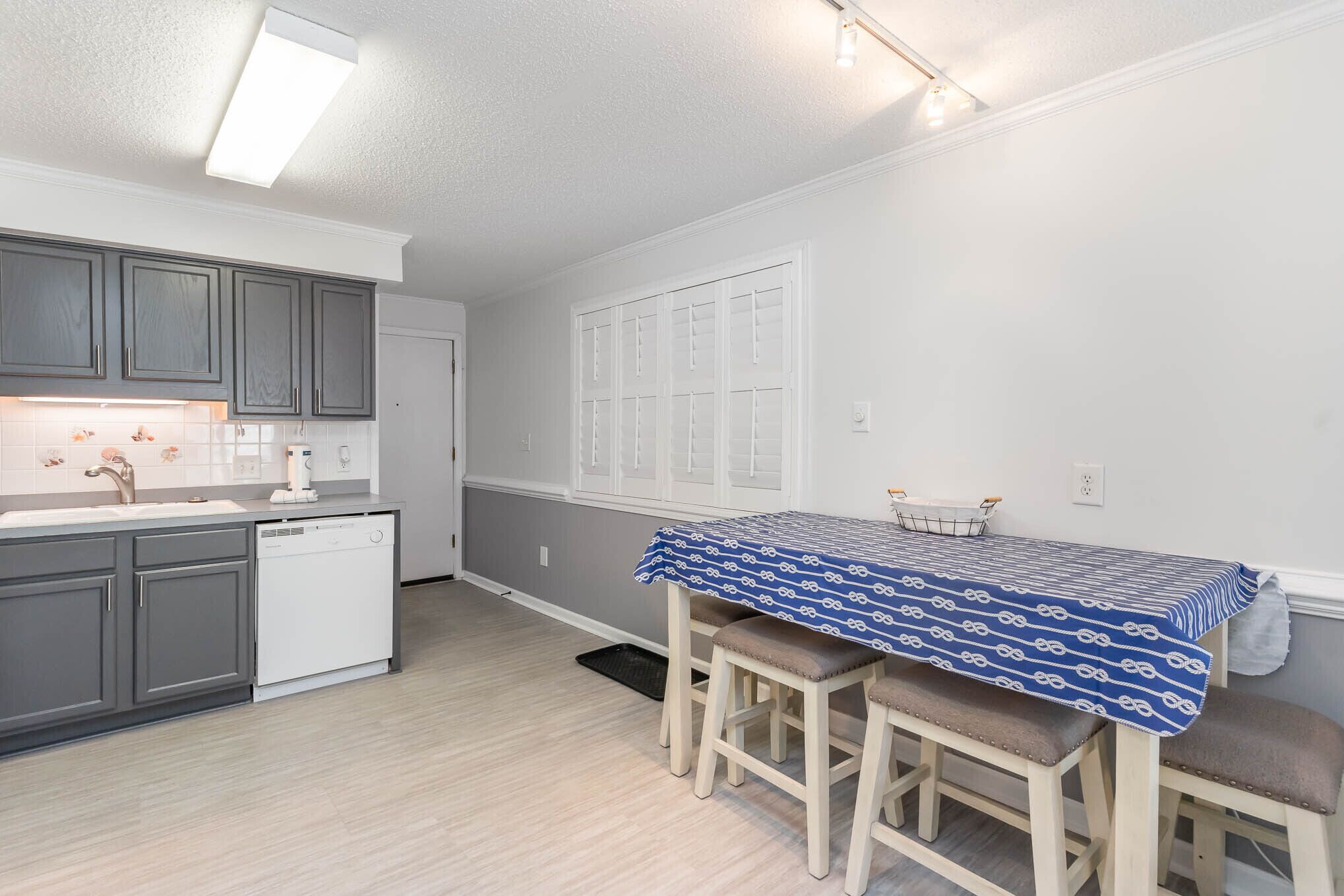 A modern kitchen with grey cabinets, a white dishwasher, and a dining area with a blue patterned tablecloth and wooden stools.