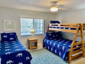 A room with two bunk beds, a small table with a lamp, a window with vertical blinds, and a ceiling fan. - Beachfront (Cape Canaveral)