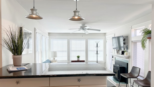 A modern kitchen with white cabinetry, a black countertop, pendant lights, a ceiling fan, and a large window with blinds. - Charming 3-Bedroom Home Near Downtown Portsmouth! (Kittery)