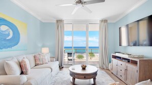A room with a ceiling fan, a sofa with cushions, a coffee table, a TV, and a view of the ocean through large windows. - 🌊Gulf Views~FREE Beach Svc~Kayak/SUP2~Pools+Splash Pad~BeachFRONT~3BR Azure 407 (Ft. Walton Beach)