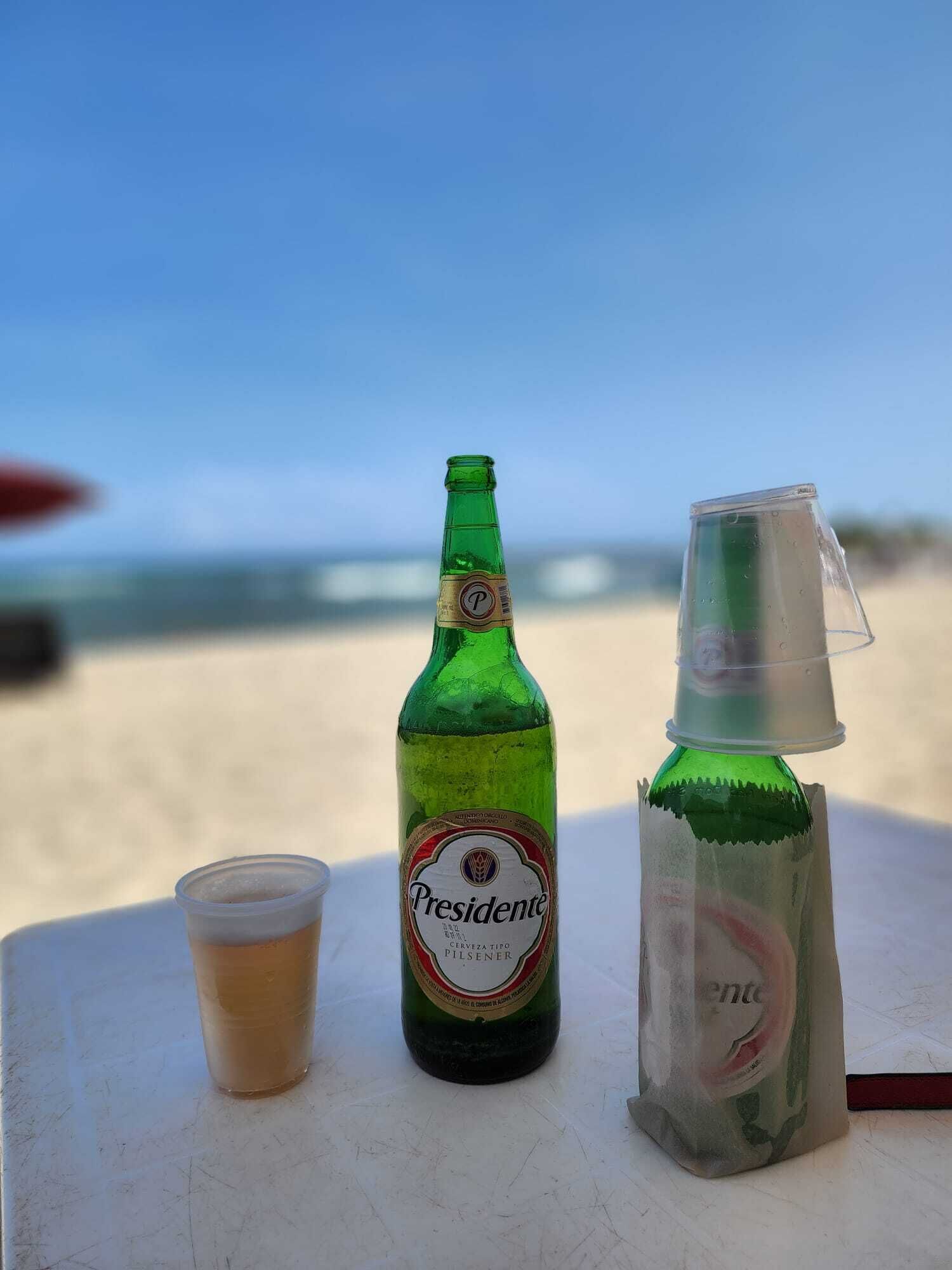 En grøn flaske med mærket 'Presidente' og et plastikbæger på et bord med strandudsigt i baggrunden.