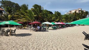 A beach with outdoor seating under umbrellas, palm trees, and a building in the background. - Modern Apartment (Juan Dolio)
