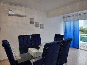 Un comedor con una mesa de vidrio y cuatro sillas tapizadas en azul. Hay un equipo de aire acondicionado en la pared y una ventana con cortinas azules.