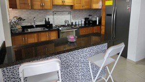 A kitchen with wooden cabinets, a stainless steel refrigerator, a gas stove, and two white bar stools. - Modern Apartment (Juan Dolio)