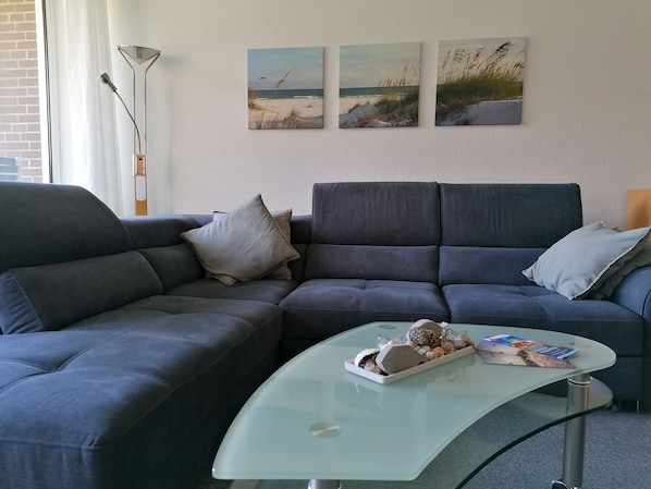 A modern living room with a dark grey sectional sofa, a glass coffee table with decorative items, and three beach-themed wall art pieces. - Schöne Wohnung in Ruhiger Lage (Borkum)