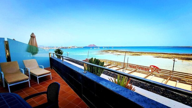 A balcony with two beige lounge chairs, a small table, and a view of the beach and ocean.