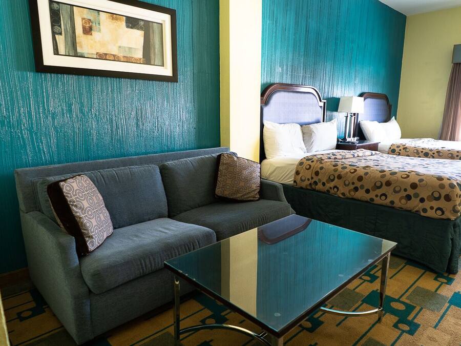 Una habitación de hotel con un sofá gris, una mesa de centro de vidrio, dos camas con ropa de cama estampada y un cuadro en la pared.