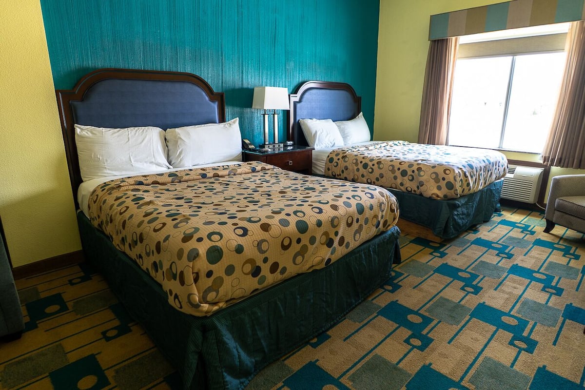 Quarto de hotel com duas camas, uma mesa de cabeceira, uma luminária e uma janela com cortinas.