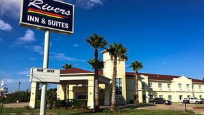 Un bâtiment de l’hôtel Rivers Inn & Suites, avec une pancarte indiquant « OUVERT MAINTENANT », des palmiers et un ciel bleu dégagé.