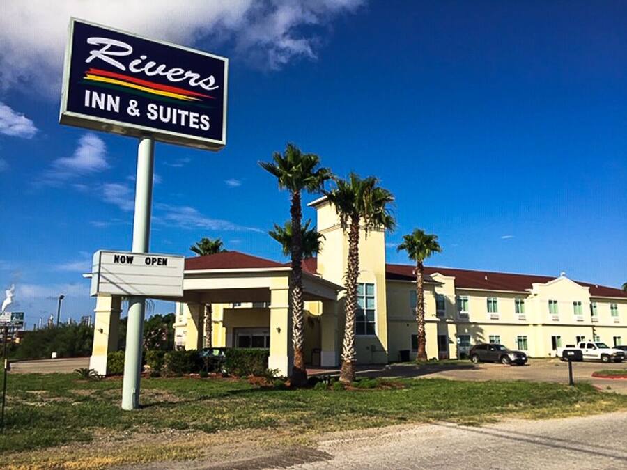 Edificio de Rivers Inn & Suites con un letrero que dice 'ABIERTO' , palmeras y un cielo azul despejado.