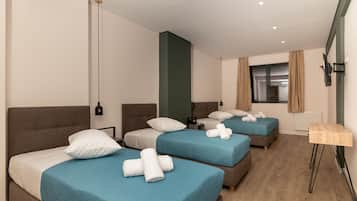 A hotel room with three beds, each with a blue bedspread and white pillows, a wooden floor, and a small table with a lamp.