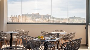 A balcony with tables and chairs, a table with breakfast items, and a view of a cityscape through large windows. - Dunlin Hotel (Athens)