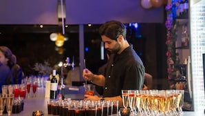 A bar area with a bartender preparing drinks. There are multiple glasses on the counter, some containing drinks. A bottle of wine and a bottle of champagne are visible. The setting appears to be indoors with a dimly lit ambiance. - Dunlin Hotel (Athens)