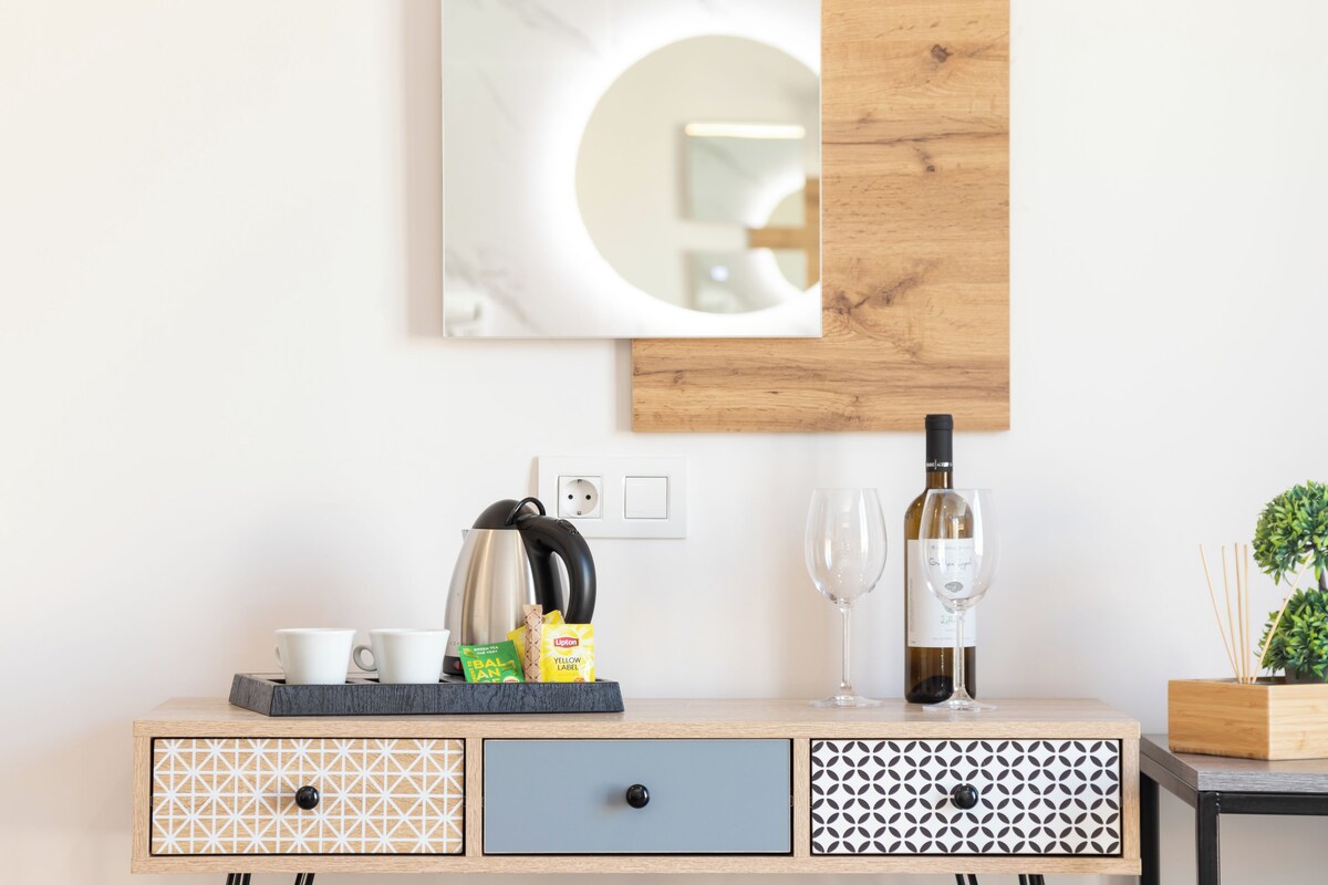 A wooden sideboard with a tray holding cups, a kettle, and snacks, a wine bottle, and glasses.
