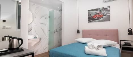 A modern hotel room with a marble bathroom, a bed with a blue quilt, a pink headboard, and a wall-mounted air conditioner.