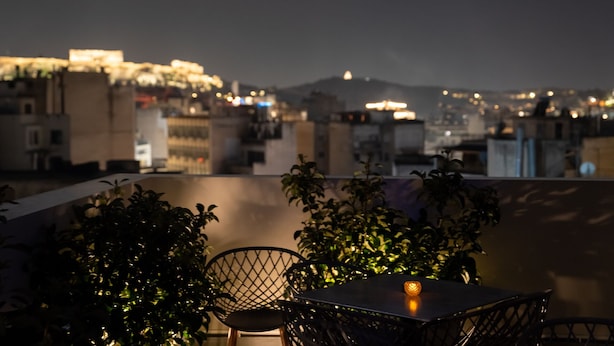 A rooftop terrace with a table and chairs, surrounded by potted plants, overlooking a cityscape at night. - Dunlin Hotel (Athens)