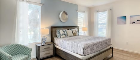 A bedroom with a bed, bedside tables, a chair, and two windows with blinds.
