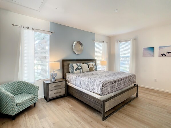 A bedroom with a bed, bedside tables, a chair, and two windows with blinds.