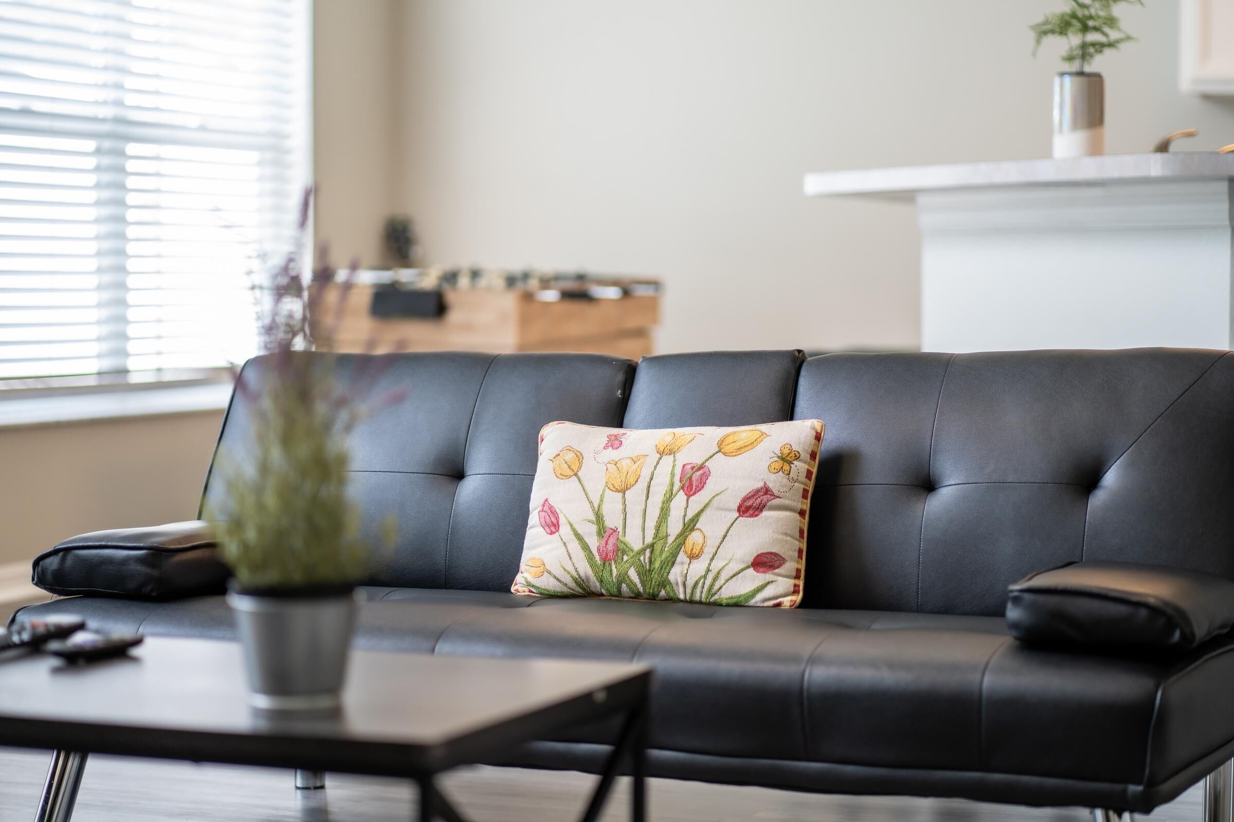 A black leather sofa with a floral pillow, a small potted plant, and a remote control on a wooden coffee table.