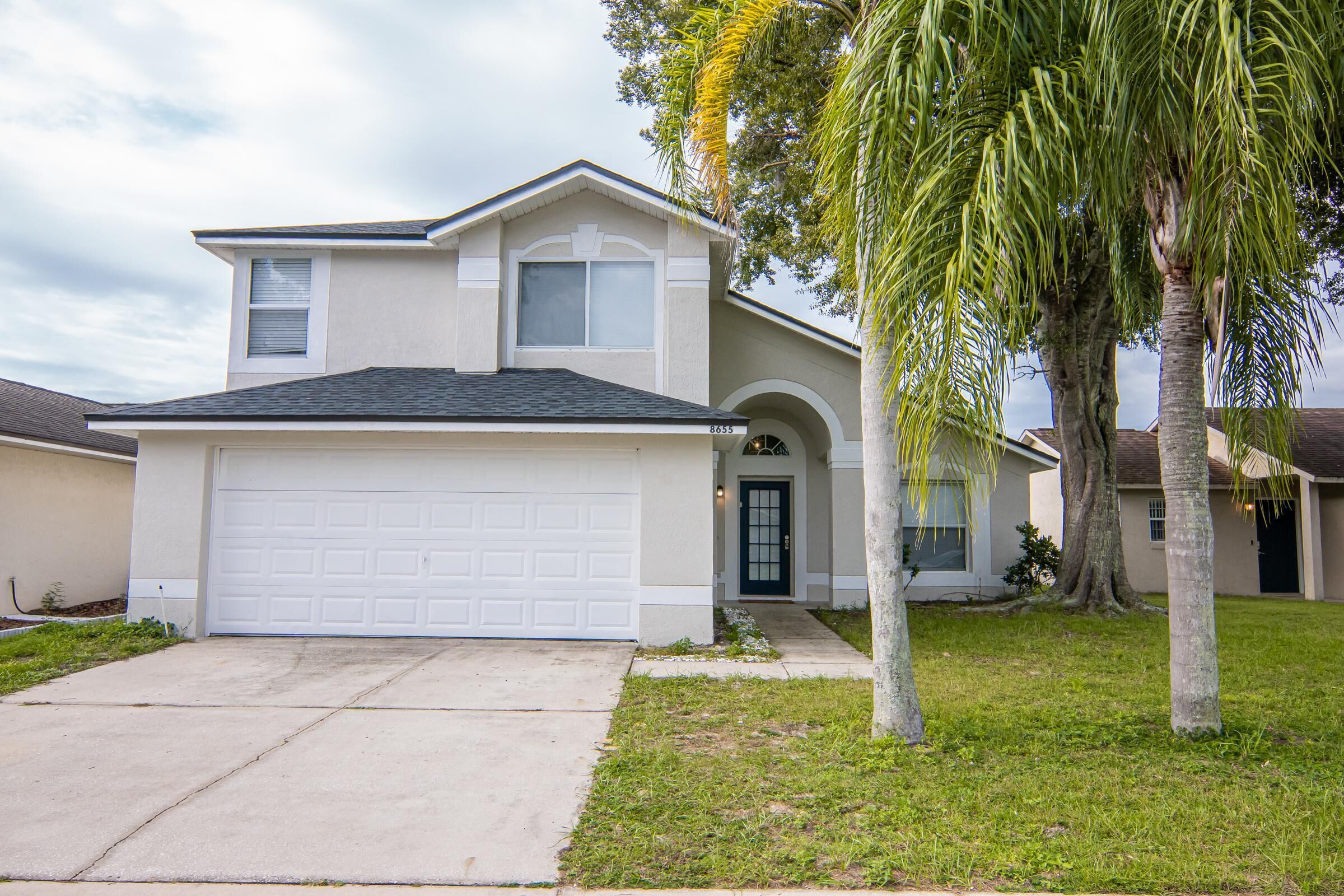 A two-story house with a garage, a front door, and a small front yard with a palm tree.
