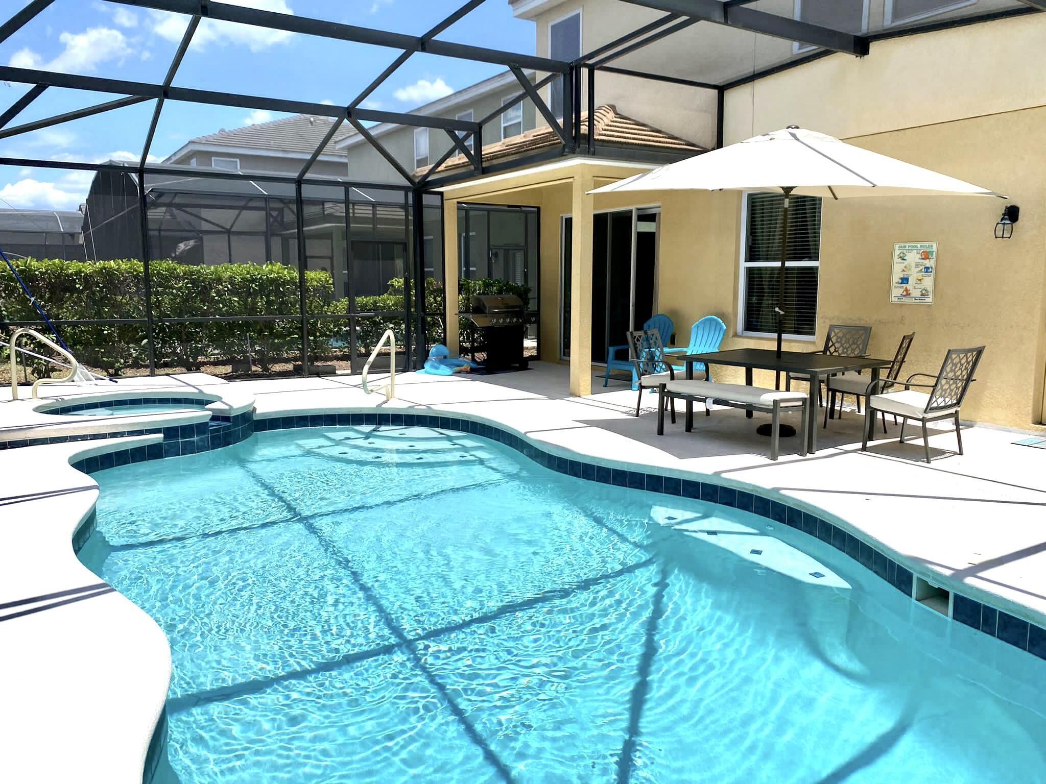 Piscina com estrutura de vidro, área de terraço com mesa e cadeiras, e vista para casas vizinhas.