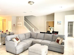 A living room with a grey sectional sofa, a small ottoman, a dining area with a table and chairs, a staircase, and a bathroom visible through an open door.