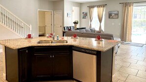 A modern kitchen with a granite countertop, built-in dishwasher, and a dining area with a gray sofa and red cups. - Solterra Resort!! Spacious House With Game Room, Private Pool and Movie Room!! (Davenport)
