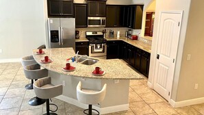 A modern kitchen with dark cabinets, stainless steel appliances, and a granite countertop. There are four bar stools and a built-in island with a sink. - Solterra Resort!! Spacious House With Game Room, Private Pool and Movie Room!! (Davenport)