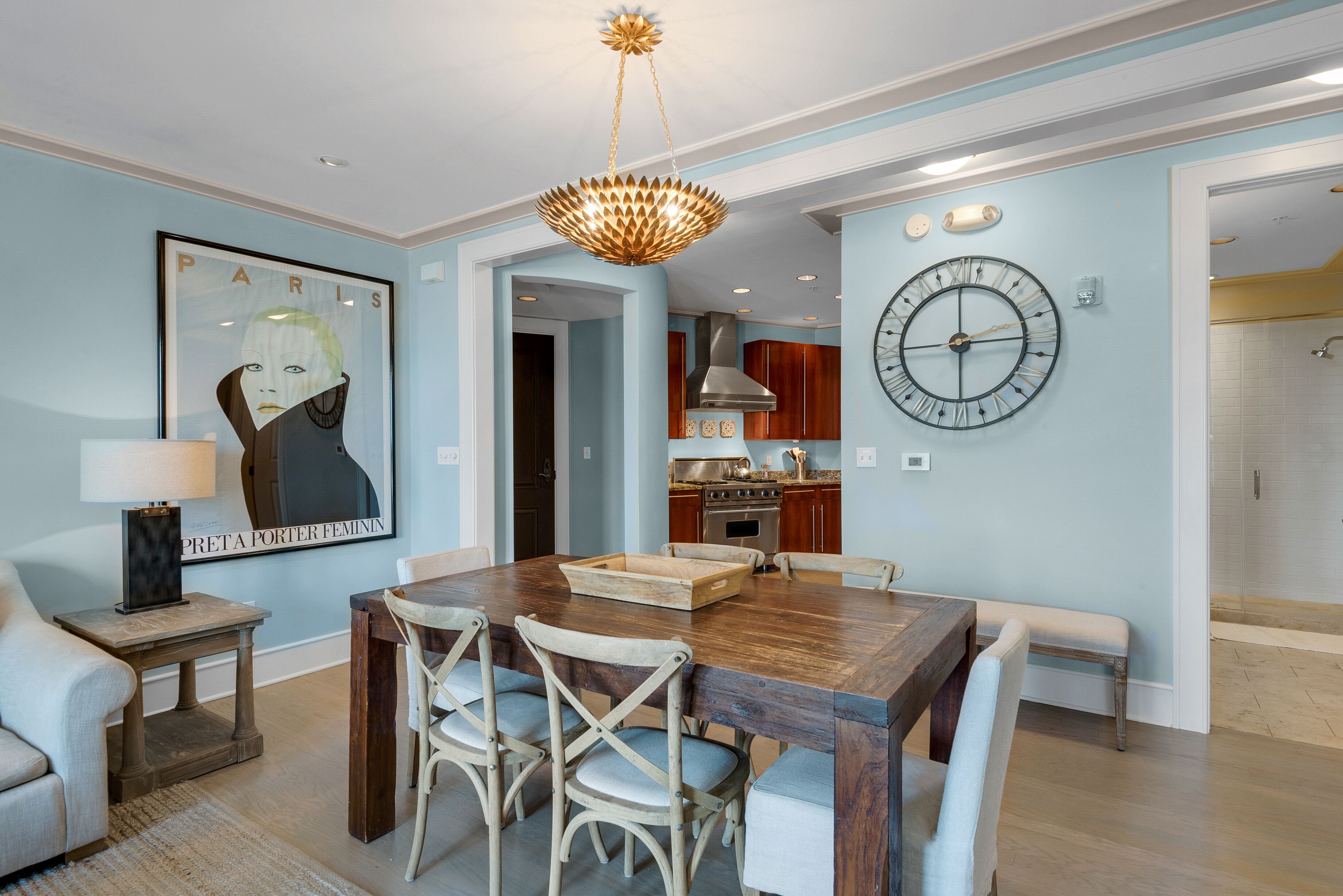 A dining area with a wooden table, four chairs, a chandelier, a clock, and a framed picture on the wall.