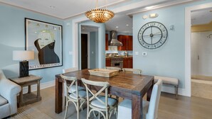 A dining area with a wooden table, four chairs, a chandelier, a clock, and a framed picture on the wall.