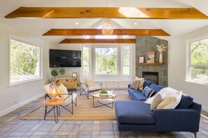 A spacious living room with a large blue sofa, wooden beams, a stone fireplace, and large windows. - Farmhouse at Sonoma Oaks. Hot Tub, Pool, Bocce! (Santa Rosa)