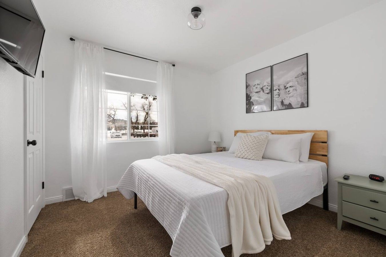 A bedroom with a bed, a nightstand, a lamp, a window with curtains, and a wall-mounted artwork.