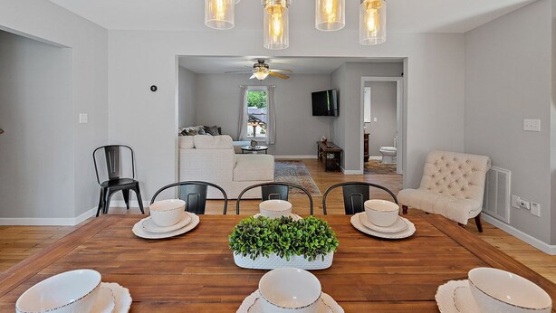 Una sala da pranzo con un tavolo in legno apparecchiato per sei persone, circondato da sedie nere. Si nota una ventola a soffitto sullo sfondo, e l'ambiente presenta colori neutri, con pareti bianche e un divano color beige.