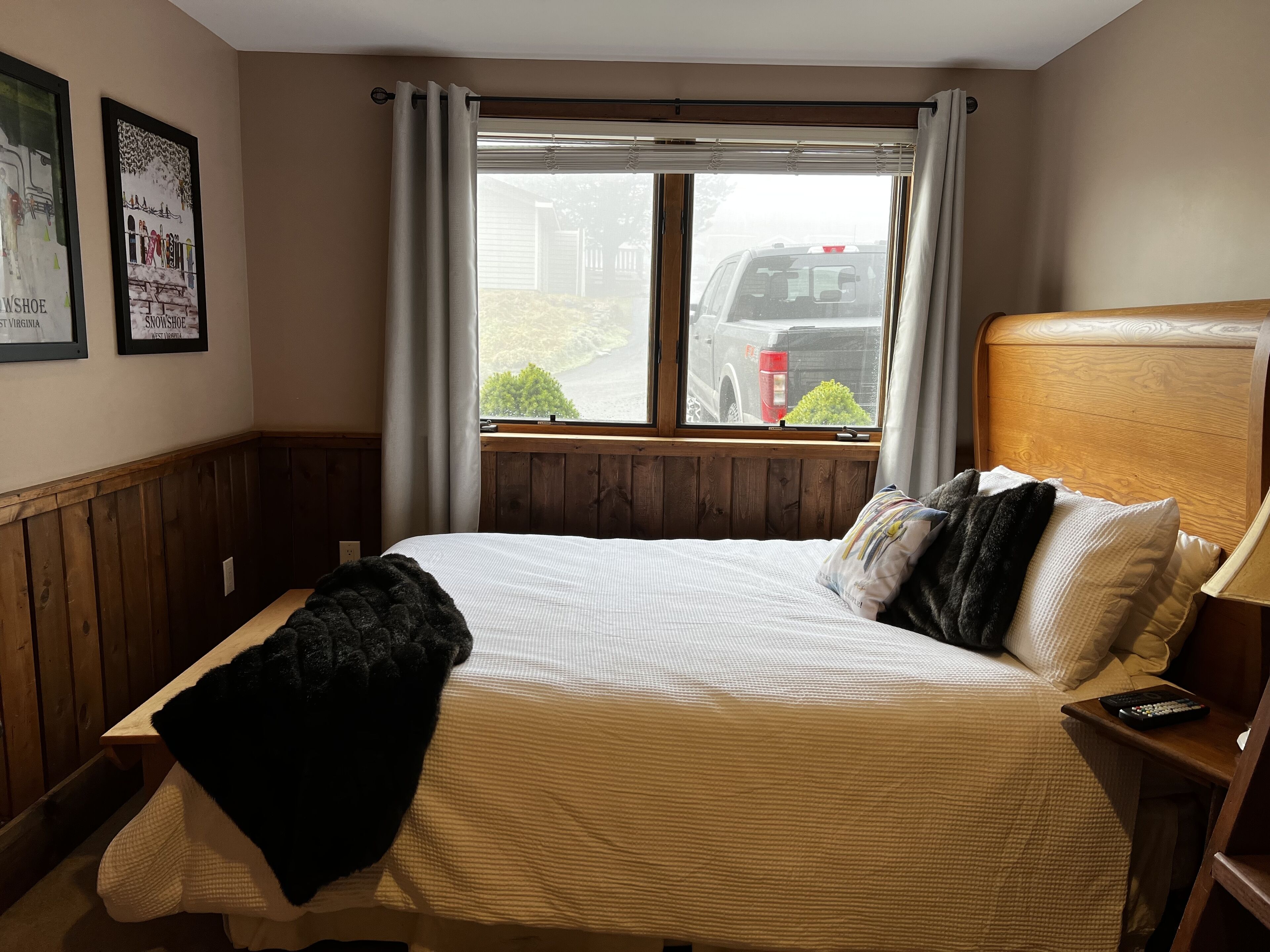 A hotel room with a large bed, wooden headboard, and a window with curtains. There are two framed pictures on the wall and a remote control on the nightstand.