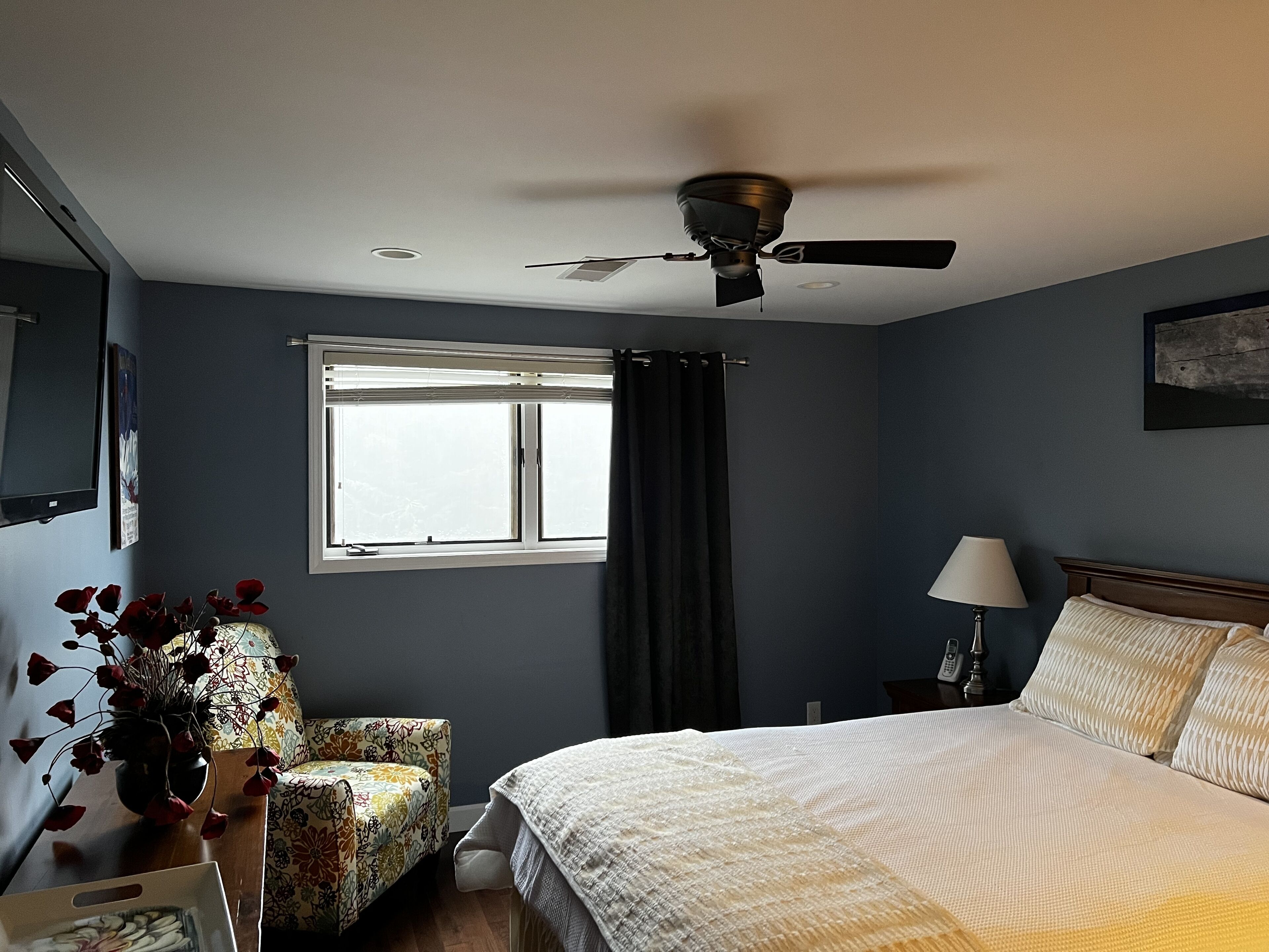 A bedroom with a bed, a chair, a window with curtains, a ceiling fan, and a nightstand with a lamp.
