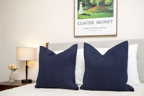 Een net opgemaakt bed met marineblauwe en witte kussens, een bedlampje en een lijst met daarin een afbeelding van Claude Monet aan de muur.