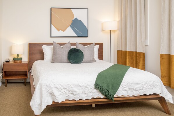 A neatly made bed with a white duvet, a green throw, and multiple pillows. There is a wooden headboard, a bedside table with a lamp, and a framed abstract artwork on the wall.